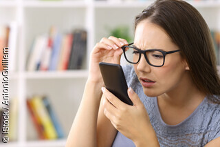 Eine junge FRau mit Brille schaut entgeistert auf ein Handy, das sie dicht vor ihr Gesicht hält.
