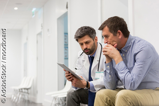 Arzt und Patient sitzen auf einem Gang und besprechen sich währenddessen sie auf ein Tablet schauen.